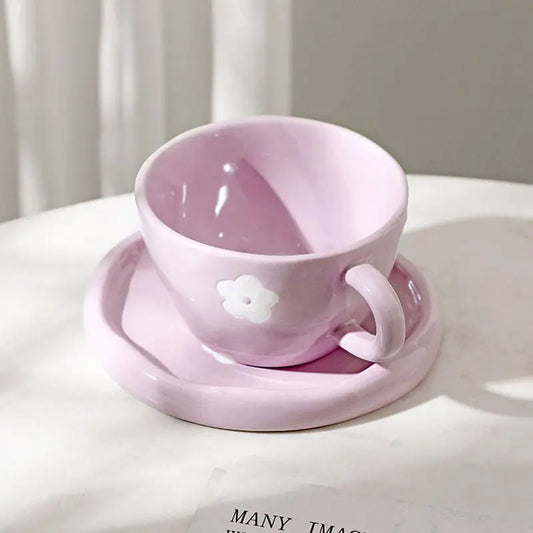 A pink floral coffee cup with a matching saucer, hand painted with purple and white flowers, displayed on a table with books and a spoon.