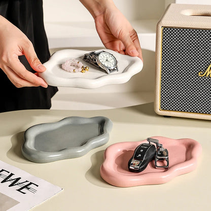 A set of three ceramic jewellery trays in white, grey, and pink, with a cloud-shaped design, displayed on a table with a person placing items like a watch and small flowers in them.